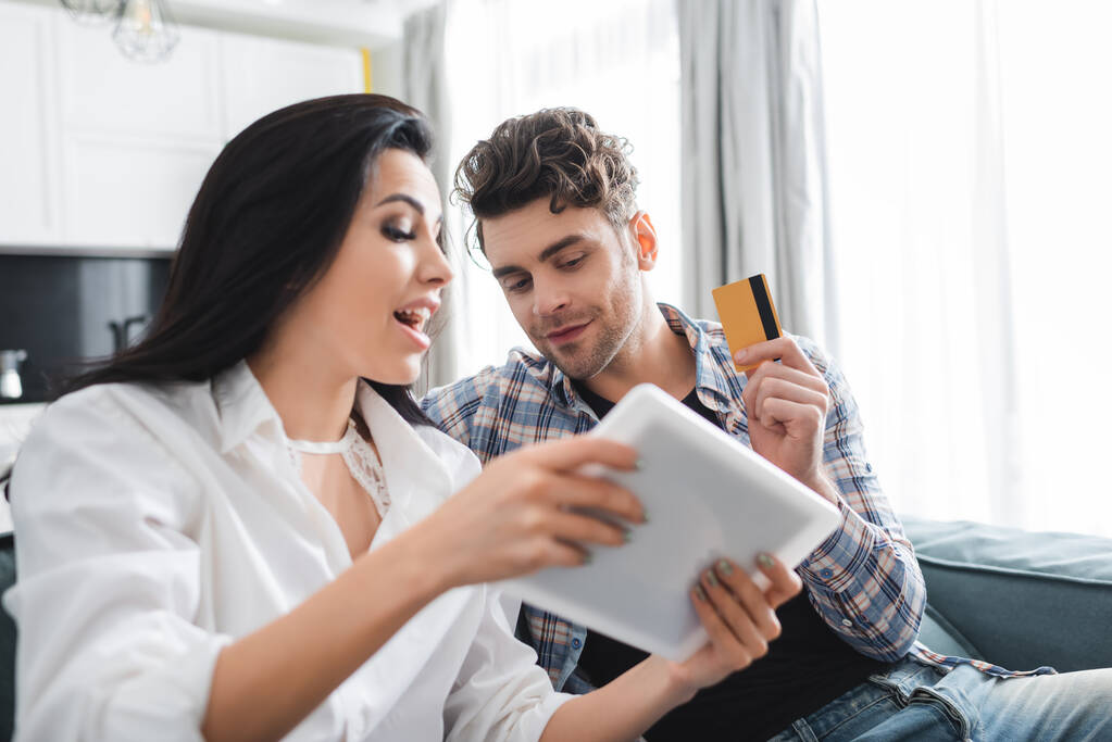 Selektivní zaměření muže držícího kreditní kartu poblíž vzrušené přítelkyně ukazující digitální tablet doma  - Fotografie, Obrázek