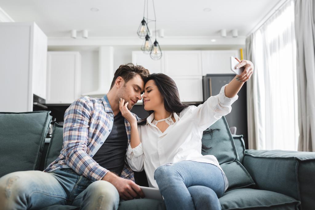 Foco seletivo da jovem mulher tocando namorado enquanto toma selfie com smartphone em casa
  - Foto, Imagem