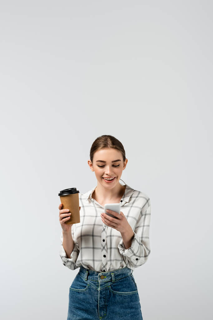 souriant attrayant fille en utilisant smartphone et tenant tasse en papier isolé sur gris - Photo, image