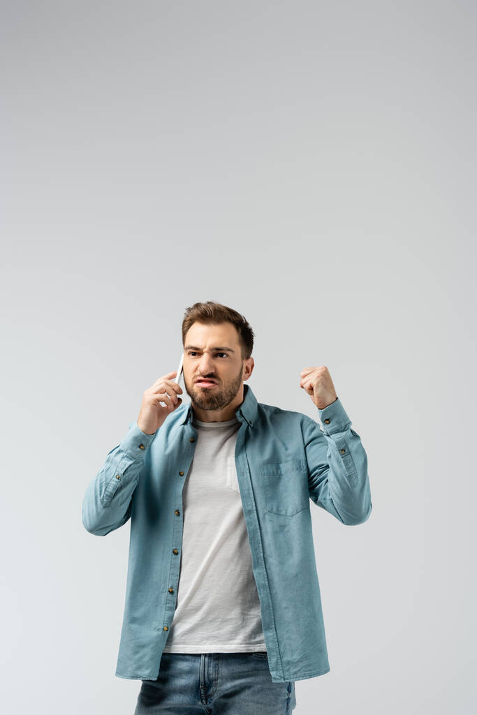 angry young man talking on smartphone and showing fist isolated on grey - Photo, Image