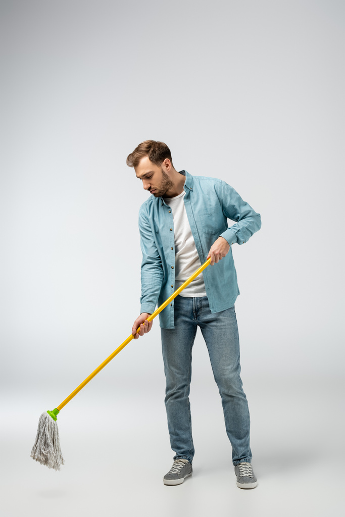 Trauriger junger Mann putzt Boden mit Wischmopp auf Grau - Foto, Bild