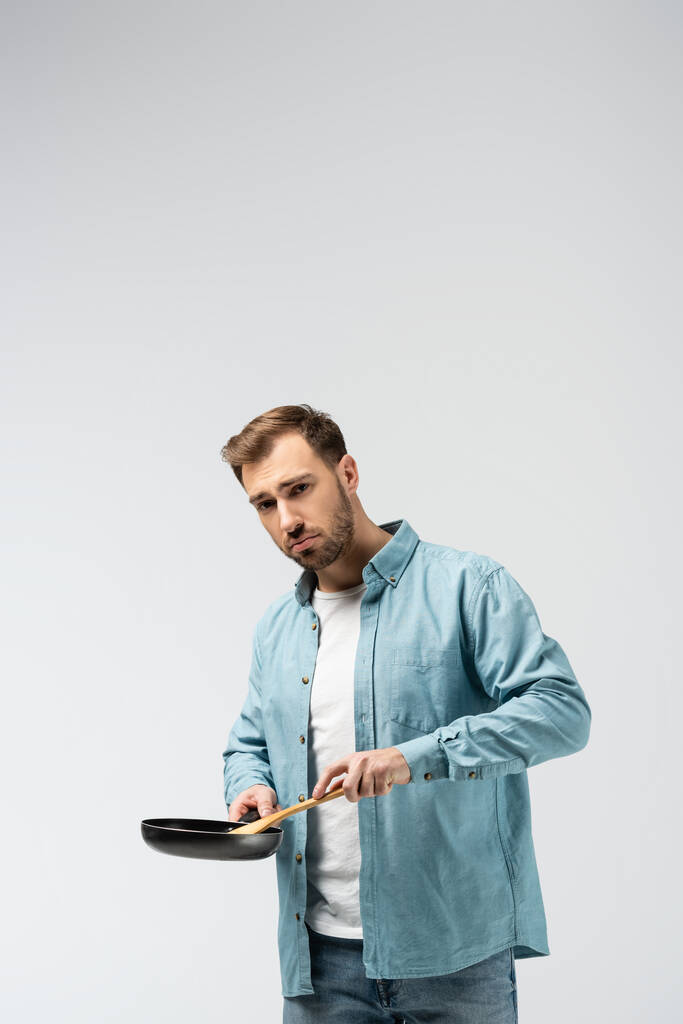 сумний молодий чоловік зі сковородою і шпателем ізольований на сірому
 - Фото, зображення