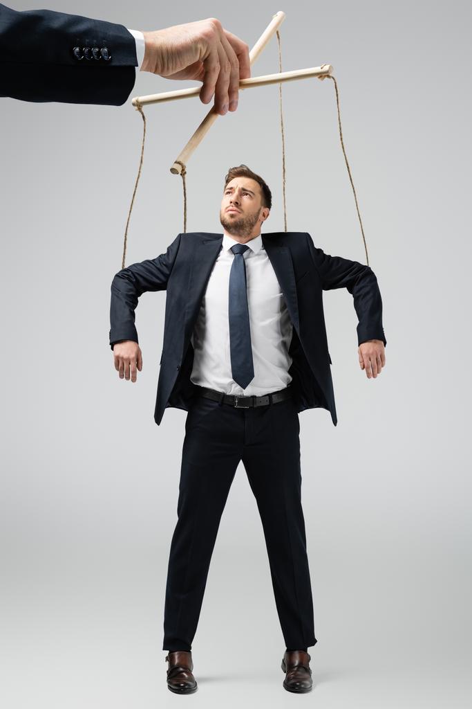vista recortada del titiritero sosteniendo marioneta de hombre de negocios en cuerdas aisladas en gris
 - Foto, Imagen