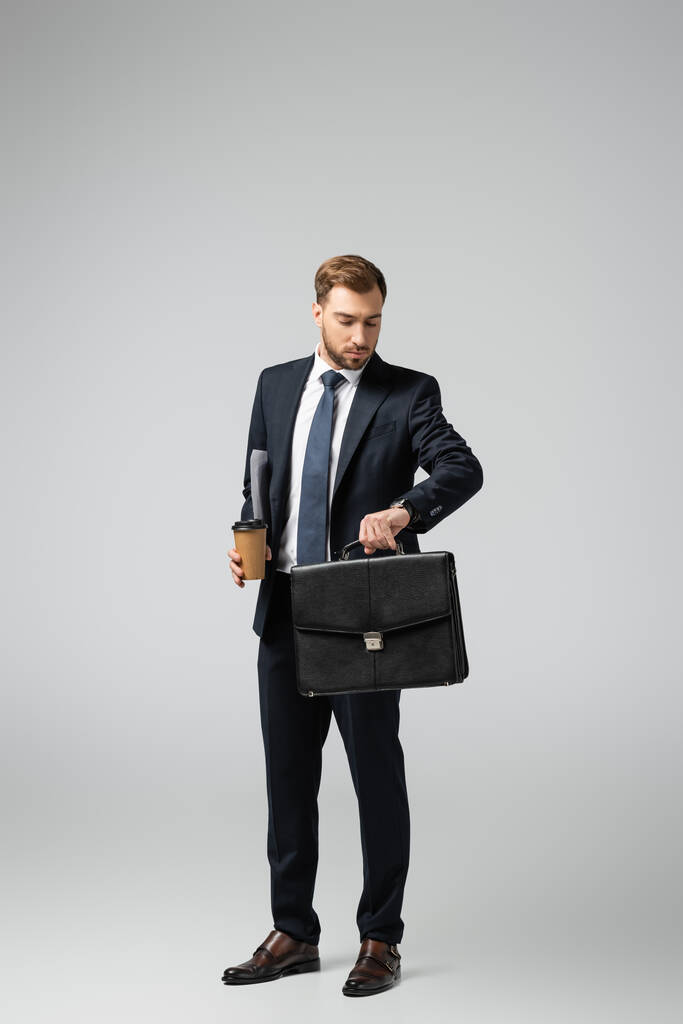 бізнесмен зі шкіряною валізою і паперовою чашкою дивиться на наручний годинник ізольовано на сірому
 - Фото, зображення