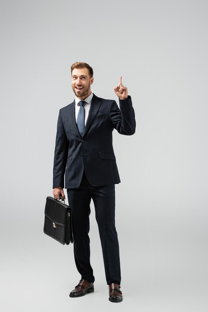 glimlachende zakenman met leren koffer naar boven gericht geïsoleerd op grijs - Foto, afbeelding