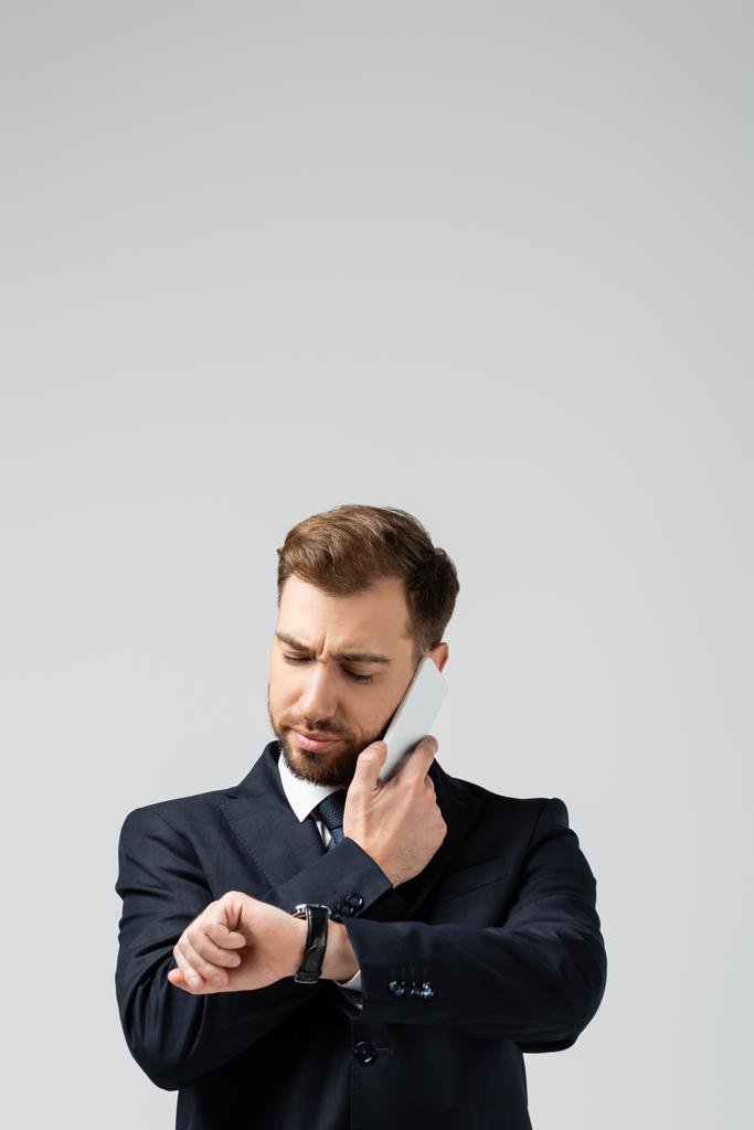 serious handsome businessman in suit talking on smartphone and looking at wristwatch isolated on grey - Photo, Image