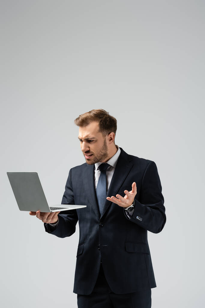 сердитый красивый бизнесмен в костюме с ноутбуком, изолированным на сером
 - Фото, изображение