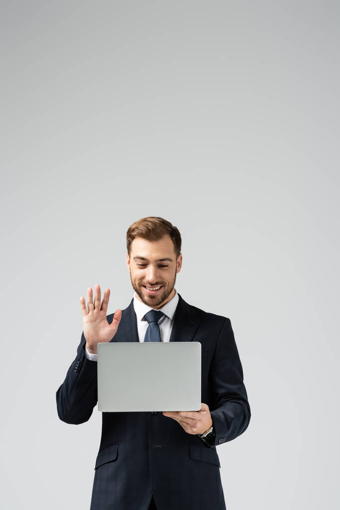 usmívající se pohledný podnikatel v obleku s video chat na notebooku izolované na šedé - Fotografie, Obrázek