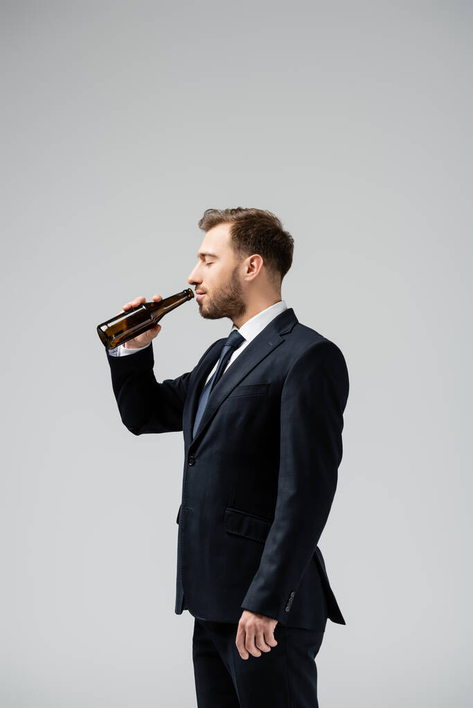 vue latérale du bel homme d'affaires en costume buvant de la bière isolée sur gris - Photo, image