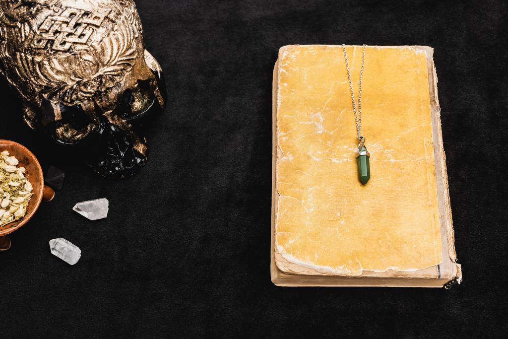 Draufsicht auf Amulett auf gealtertem Buch in der Nähe von Sagebrush, okkultem Voodoo-Schädel und Kristallen auf Schwarz - Foto, Bild