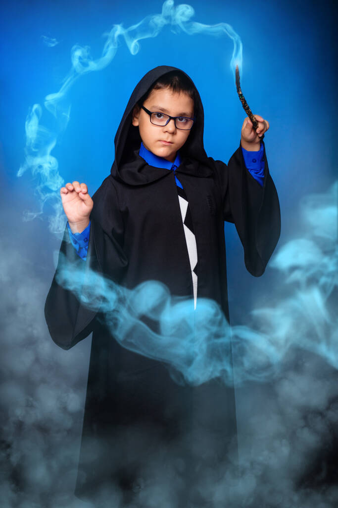 Un niño mago en gafas con una varita mágica en sus manos y con una bata negra con una capucha evoca un hechizo mientras está de pie en la niebla y el humo
. - Foto, imagen