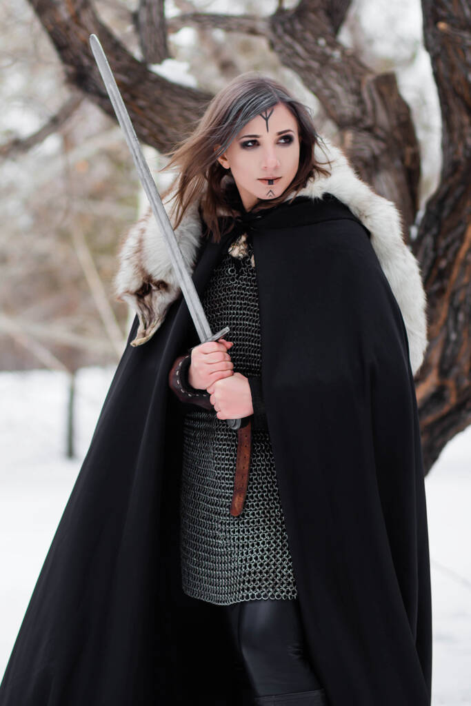  Portré egy középkori nőről, a Viking kor harcosáról a téli erdőben, egy nagy fa hátterében. Láncba verve, köpenyben, bundában, bőrnadrágban és csizmában, karddal a kezükben. Harci festék az arcon.. - Fotó, kép