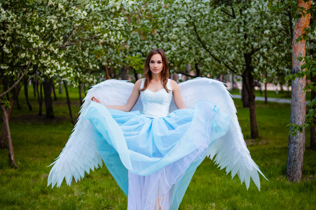 Una joven y hermosa mujer con un corsé blanco y un exuberante vestido azul con grandes alas de ángel blanco detrás de su espalda posando sobre el telón de fondo de manzanos florecientes en el parque. Ángel en el Jardín del Edén
. - Foto, imagen