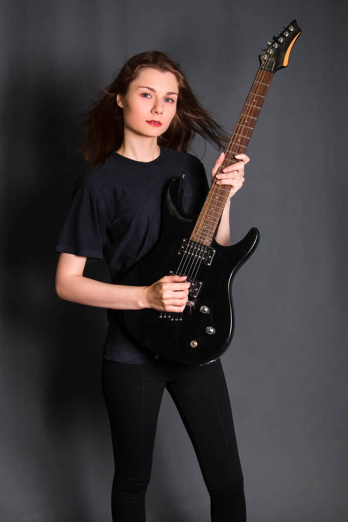 Portré egy gyönyörű, fiatal, fekete ruhás rocknőről elektromos gitárral a kezében. Stúdiókép szürke háttérrel. Tiszta bőrű modell. - Fotó, kép