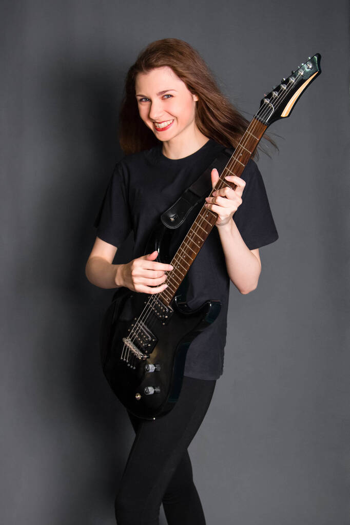 Portret van een mooie, jonge rockvrouw in zwarte kleren met een elektrische gitaar in haar handen. Studio foto op een grijze achtergrond. Model met schone huid. - Foto, afbeelding