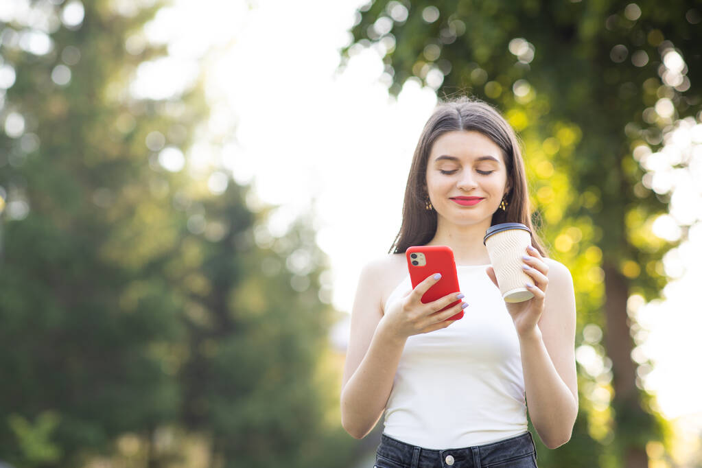Dziewczyna śmieje się patrząc na swój smartfon. Krajobraz miejski z drzewami. Wesoła młoda dziewczyna z papierową filiżanką kawy w lewej ręce - Zdjęcie, obraz