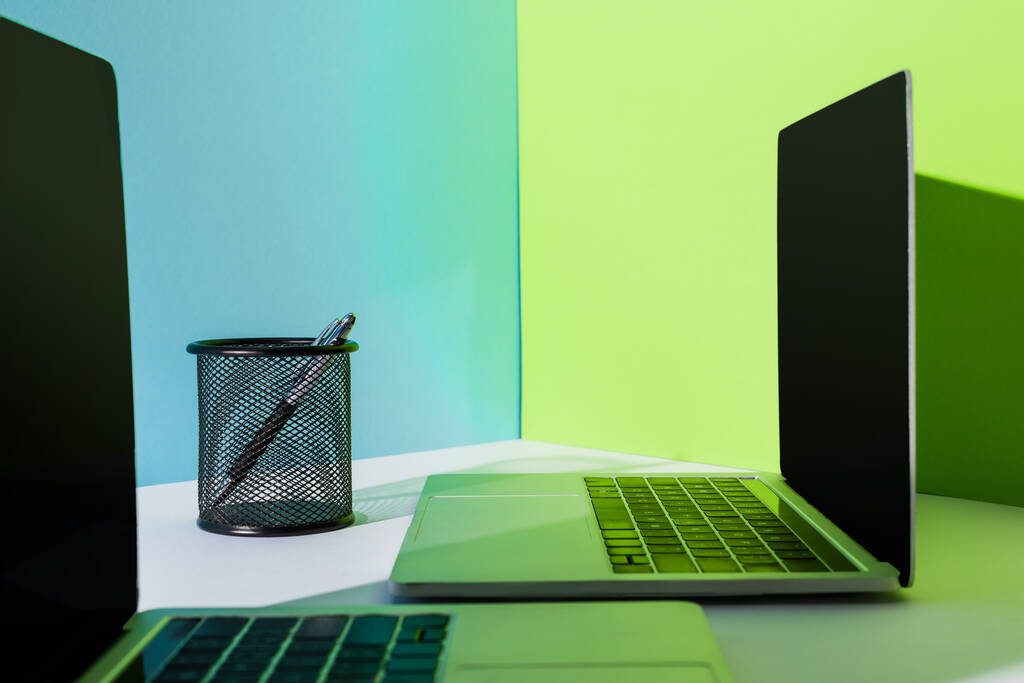 pen in houder bij moderne laptops op blauwe, witte en groene achtergrond - Foto, afbeelding