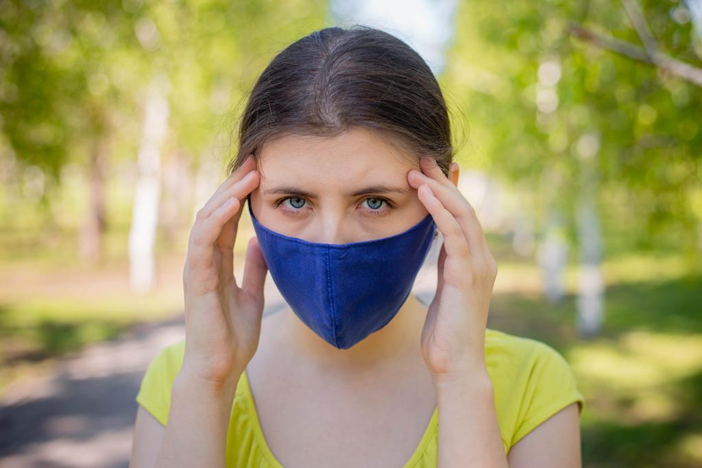 Eine junge Frau in blauer Maske hält ihren Kopf in den Händen. Das Konzept von Kopfschmerzen und schlechter Gesundheit aufgrund der Selbstisolation während der Coronavirus-Pandemie.    - Foto, Bild