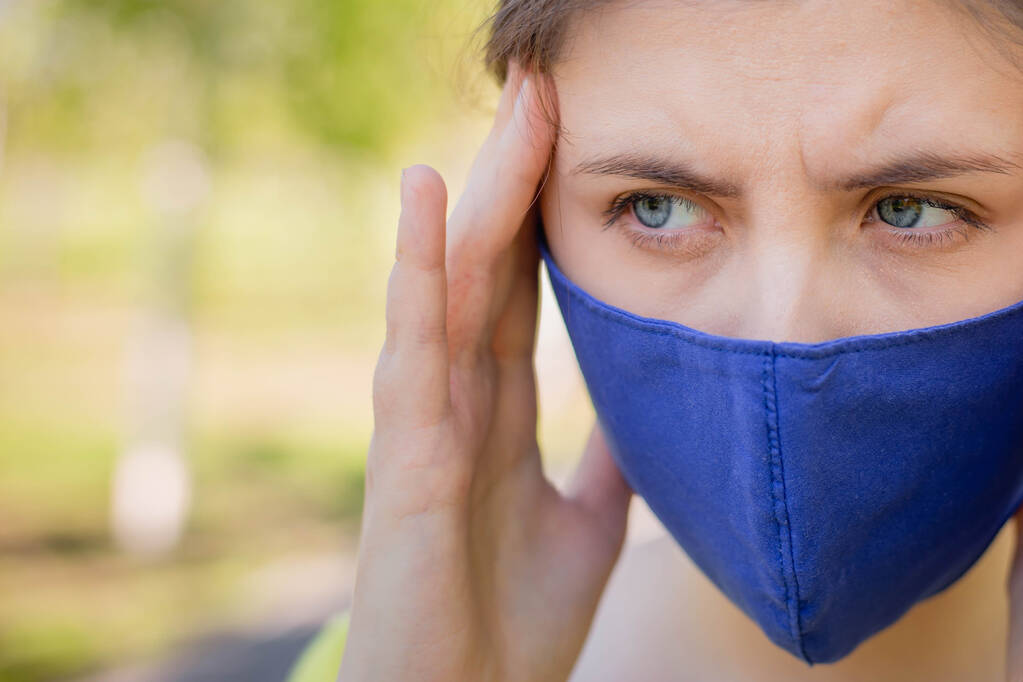 Una joven con una máscara azul sostiene su cabeza en sus manos. El concepto de cefalea y mala salud debido al autoaislamiento durante la pandemia de coronavirus. Copiar espacio
.  - Foto, Imagen