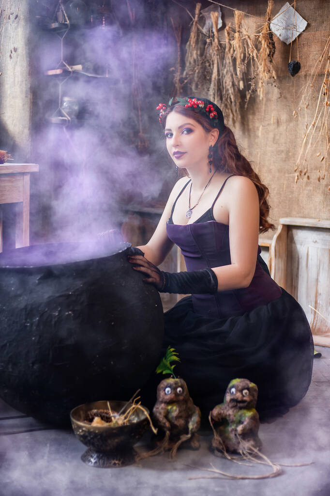 Młoda, atrakcyjna czarownica warzy eliksir w dużym czarnym kotle emitującym magiczny fioletowy dym. - Zdjęcie, obraz