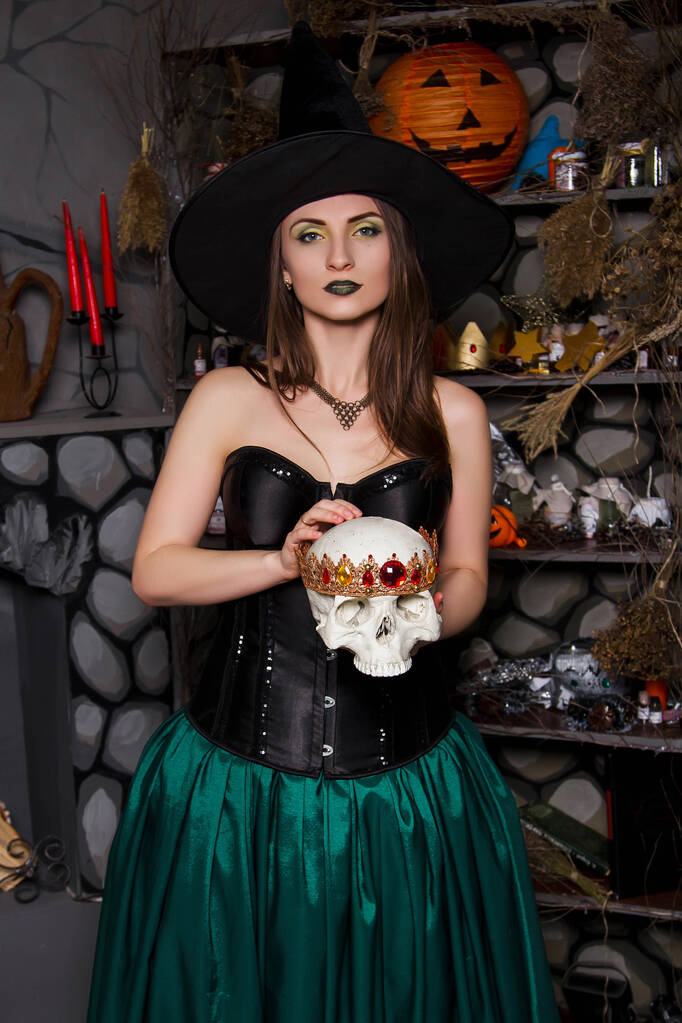 Nuori, kaunis nainen vihreässä hameessa, musta korsetti ja musta hattu pitelevät kruunua käsissään. Noitapuku, Halloween, valokuvaamo, malli puhtaalla iholla. - Valokuva, kuva