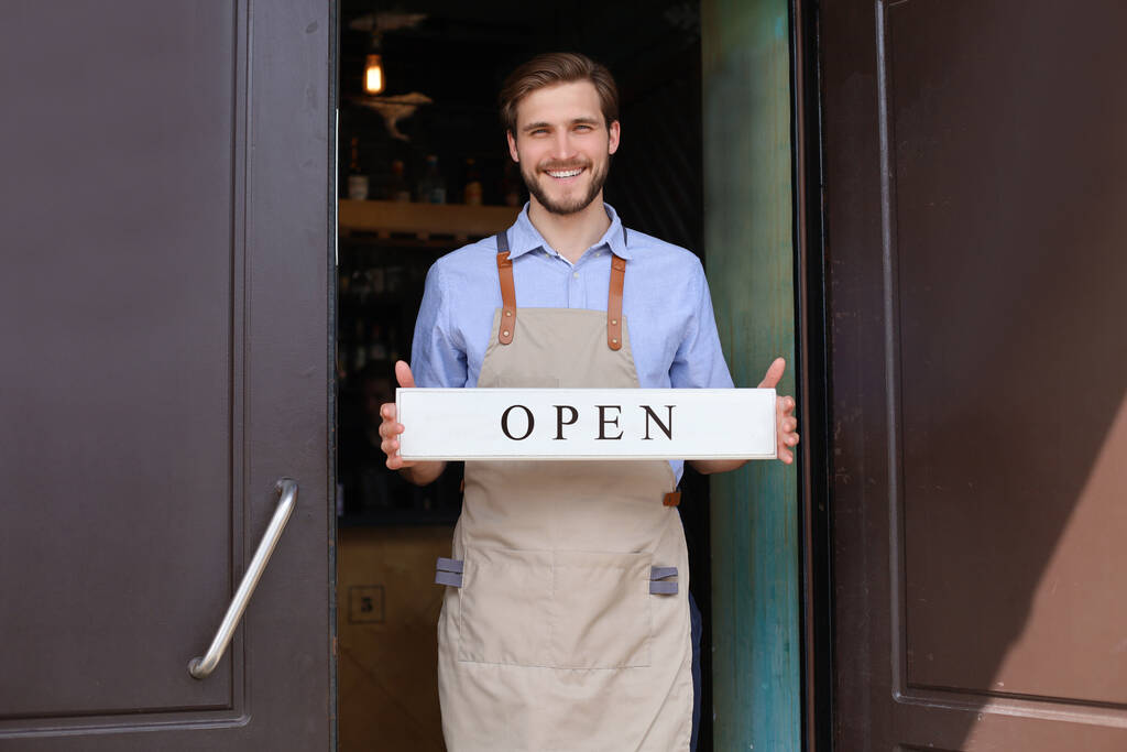 Smiling male owner of coffee shop is standing outdoors. He is holding placard and inviting everybody to come in. Open - Photo, Image