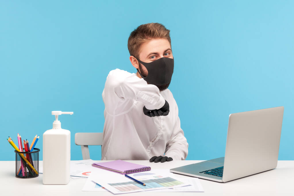 Pozitivní kancelářský pracovník, který zdraví narážející loktem do kamery, nosí hygienickou masku a ochranné rukavice, aby zabránil vzniku koronaviru na pracovišti, koncepci zdravotní péče, pandemii covid-19. studio pořízeno izolovaně - Fotografie, Obrázek