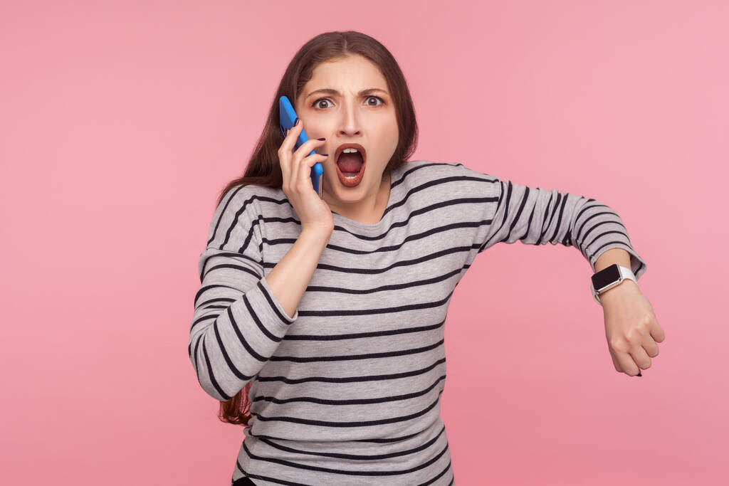 Porträt einer ungeduldigen Frau in gestreiftem Sweatshirt, die mit einem Handy spricht und mit schockiertem, wütendem Gesichtsausdruck schaut, die Armbanduhr zeigt, verrückt nach der späten Zeit. Studioaufnahme isoliert auf rosa Hintergrund - Foto, Bild