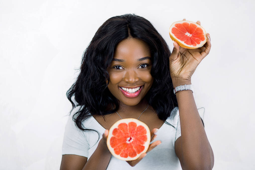 Kesä, hedelmät ja ihmiset käsite. Kauneus lähikuva muotokuva melko hymyilevä nuori afrikkalainen nainen yllään valkoinen t-paita, tilalla greippi osia käsissä, seisoo eristetty valkoisella pohjalla - Valokuva, kuva
