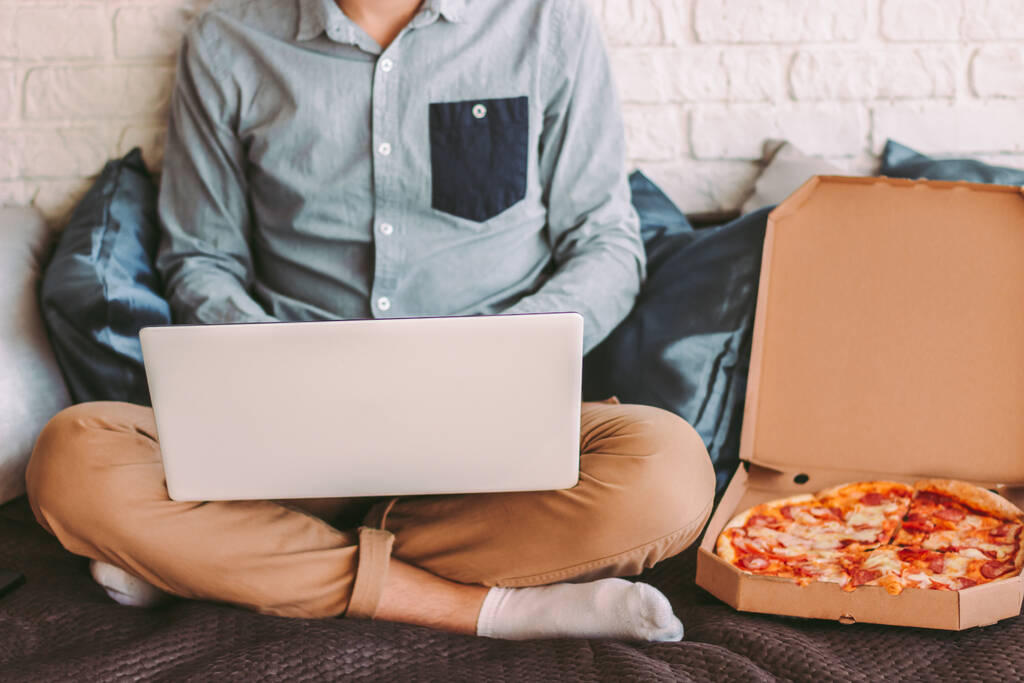 Elegante hombre hipster estudiante navegar por Internet en el ordenador portátil y comer pizza en el acogedor lugar de trabajo en el sofá. Empresario ocupado usando la computadora para el trabajo remoto en la oficina en casa. Sabrosa comida rápida para llevar para el almuerzo
 - Foto, imagen