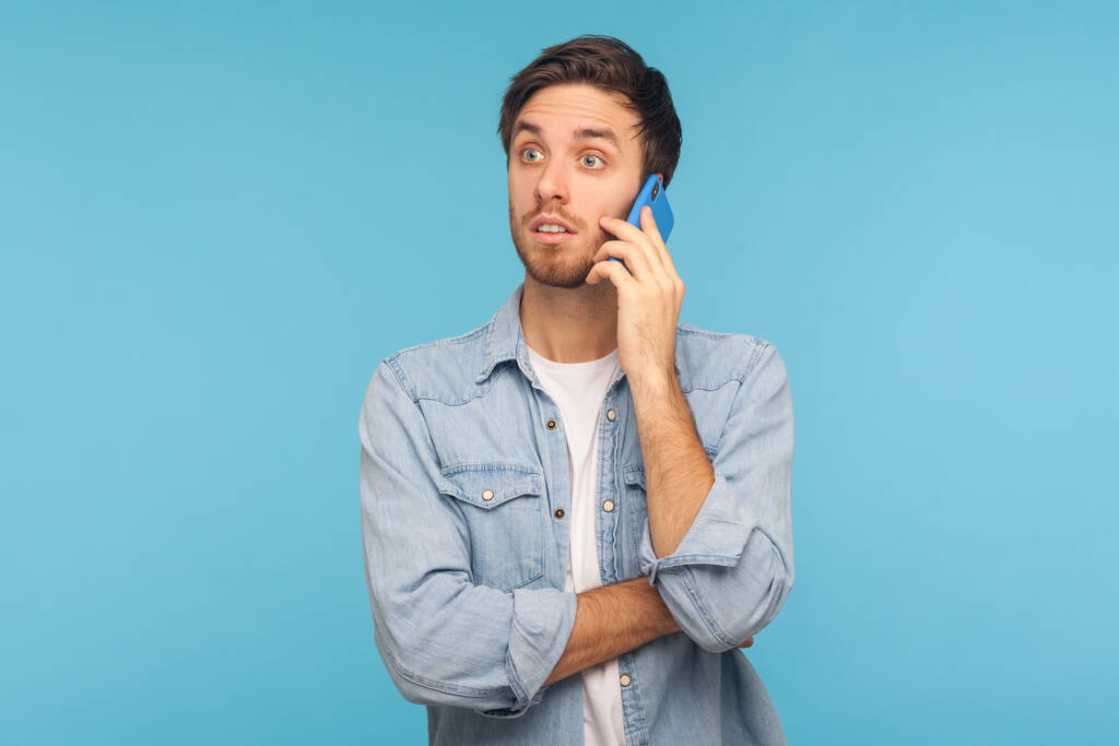 Portrait of amazed young businessman in casual denim shirt talking on phone and looking shocked, surprised by unexpected mobile conversation with partner. studio shot isolated on blue background - Photo, Image