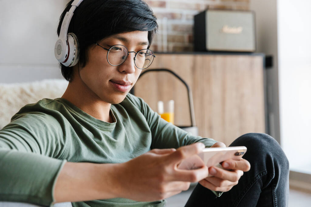 Beeld van knappe jonge aziatische man het dragen van koptelefoon met behulp van mobiele telefoon tijdens het rusten in appartement - Foto, afbeelding