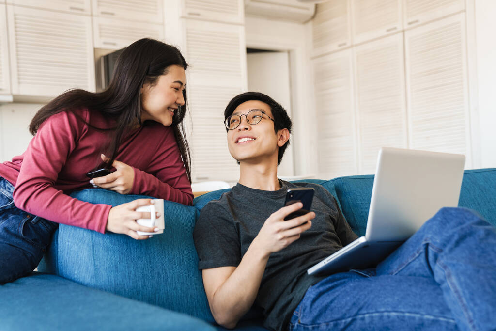 Foto de pareja asiática sonriente usando teléfono inteligente y portátil mientras bebe café en el sofá en casa
 - Foto, Imagen