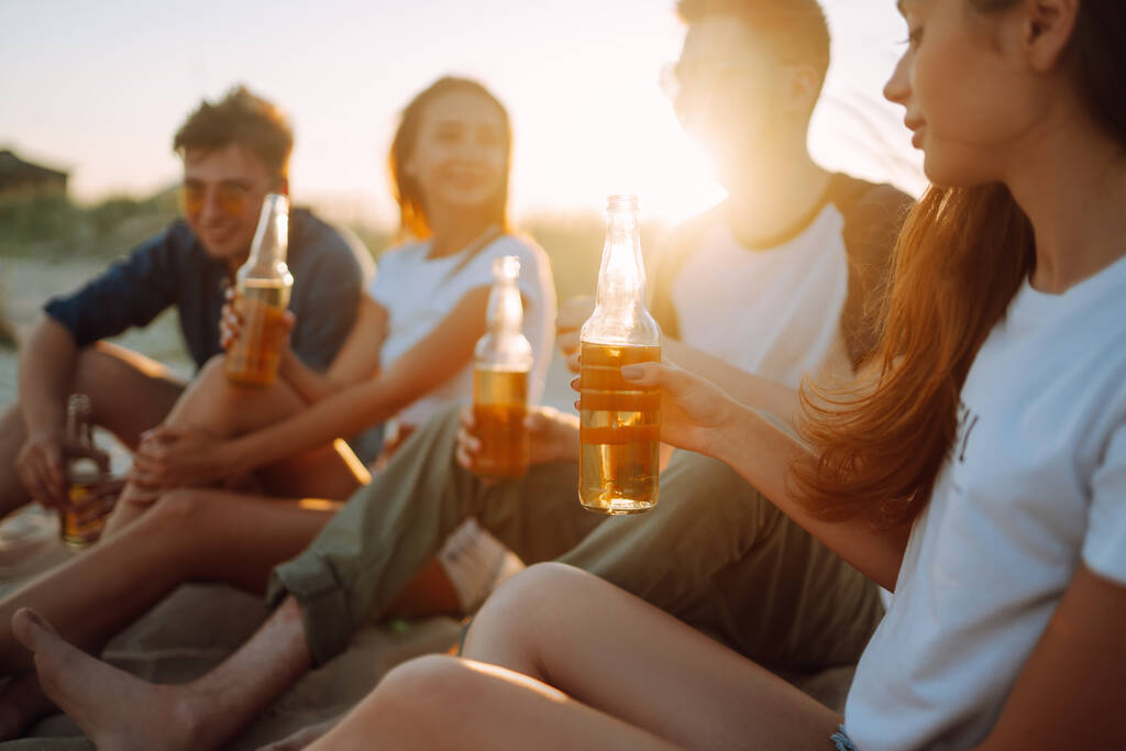 Jóvenes sentados juntos en la playa, bebiendo cerveza y dando una fiesta. Grupo de amigos aplauden con cervezas en la playa durante las vacaciones de verano
. - Foto, Imagen