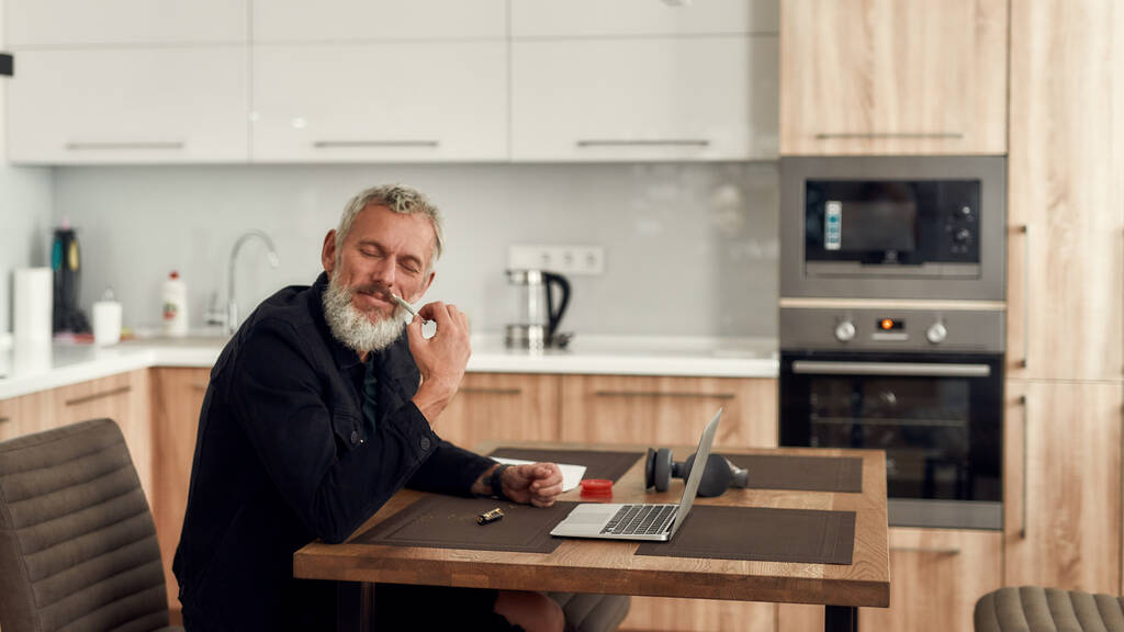 No Drama. Middle-aged man, artist enjoying the smell of marijuana cigarette or joint, sitting at home, in the kitchen and using laptop. Marijuana grinder, lighter and headphones on the table - Photo, Image