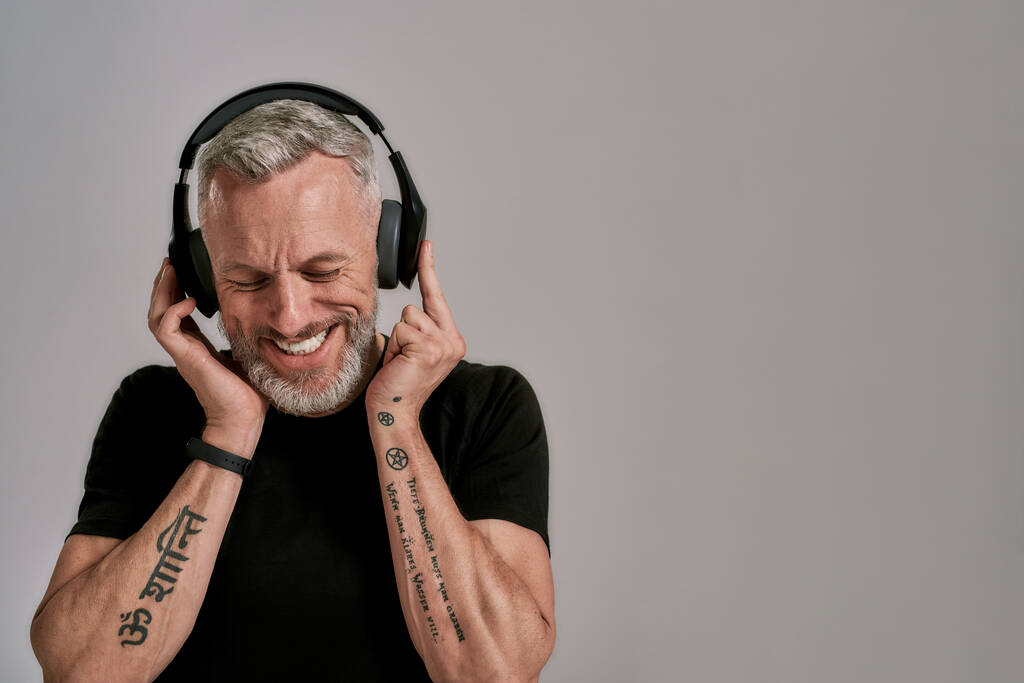 Disfruta el ritmo. Hombre musculoso de mediana edad con camiseta negra y auriculares sonriendo mientras escucha música, posando en el estudio sobre fondo gris
 - Foto, Imagen