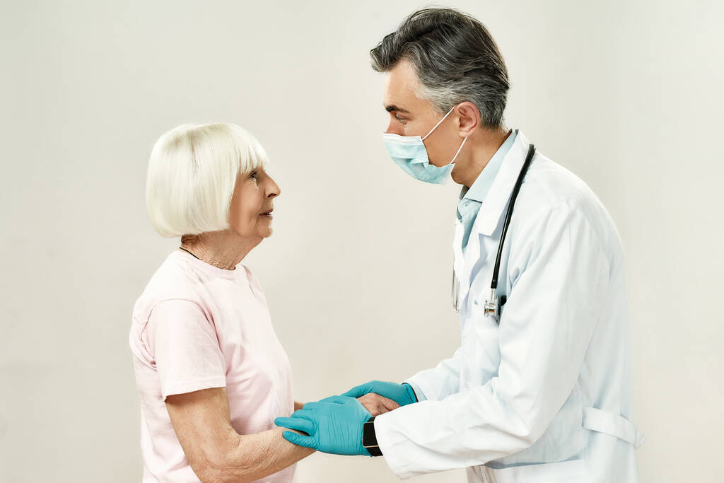 Medische zorg en ondersteuning. Professionele volwassen mannelijke arts in medisch uniform en beschermend masker hand in hand van vrouwelijke senior patiënt - Foto, afbeelding