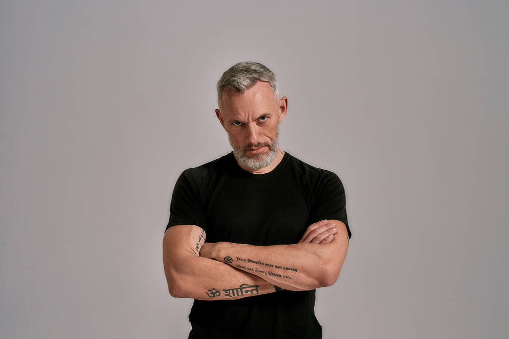Fuerza. Hombre de mediana edad con brazos cruzados en camiseta negra mirando a la cámara mientras posa en el estudio sobre fondo gris
 - Foto, imagen