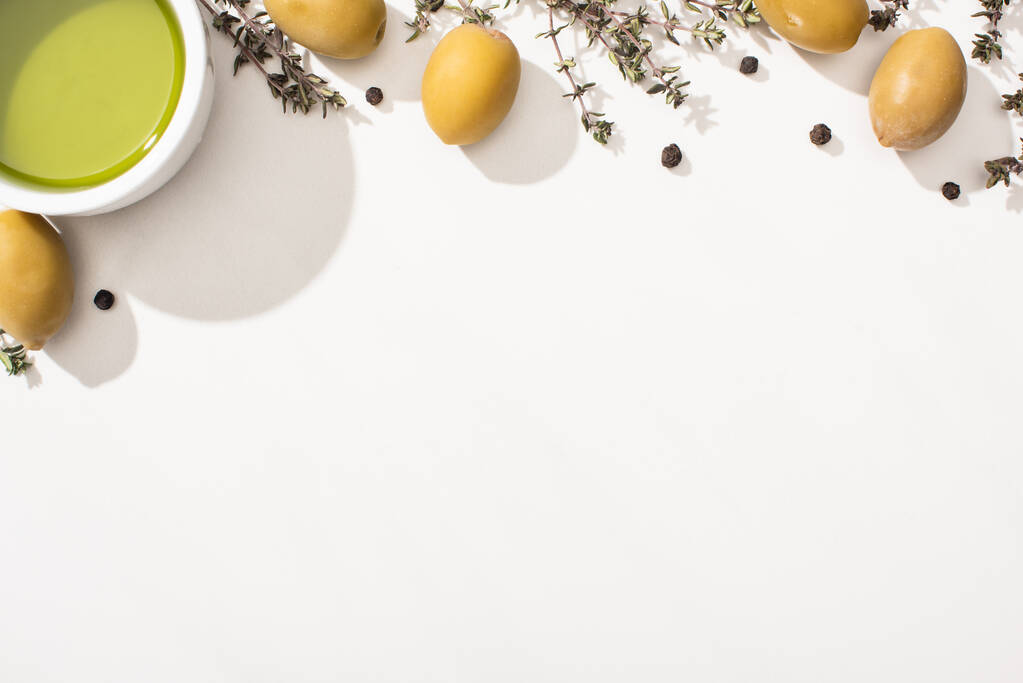вид на оливковое масло в миске рядом с зелеными оливками, травами и черным перцем на белом фоне
 - Фото, изображение