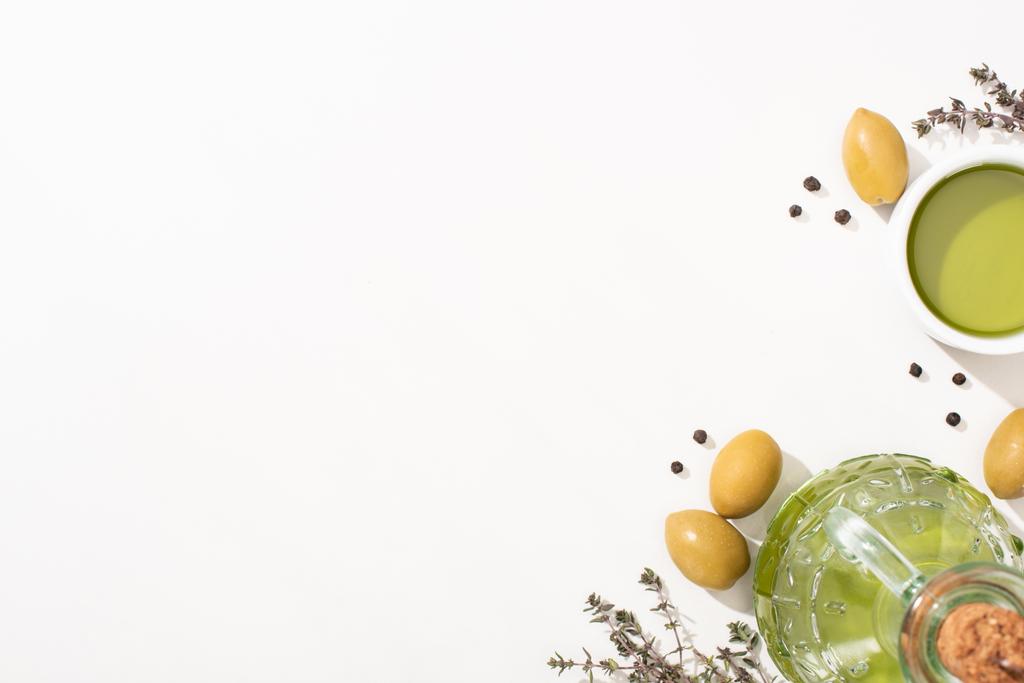 vrchní pohled na olivový olej v misce a láhev v blízkosti zelených oliv, bylina a černý pepř na bílém pozadí - Fotografie, Obrázek