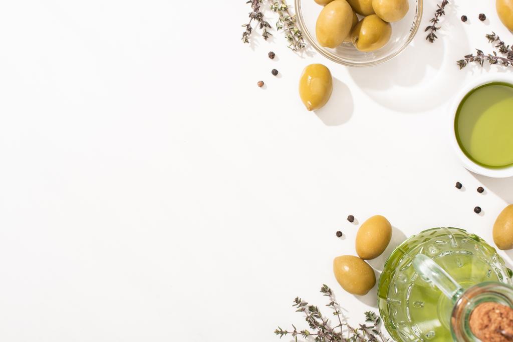 πάνω όψη του ελαιολάδου σε γυάλινο μπολ και μπουκάλι κοντά σε πράσινες ελιές, βότανο και μαύρο πιπέρι σε λευκό φόντο - Φωτογραφία, εικόνα