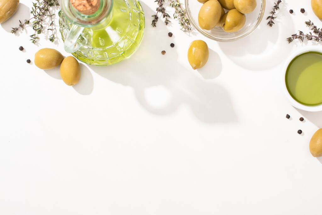 top view of olive oil in glass bowl and bottle near green olives, herb and black pepper on white background - Photo, Image