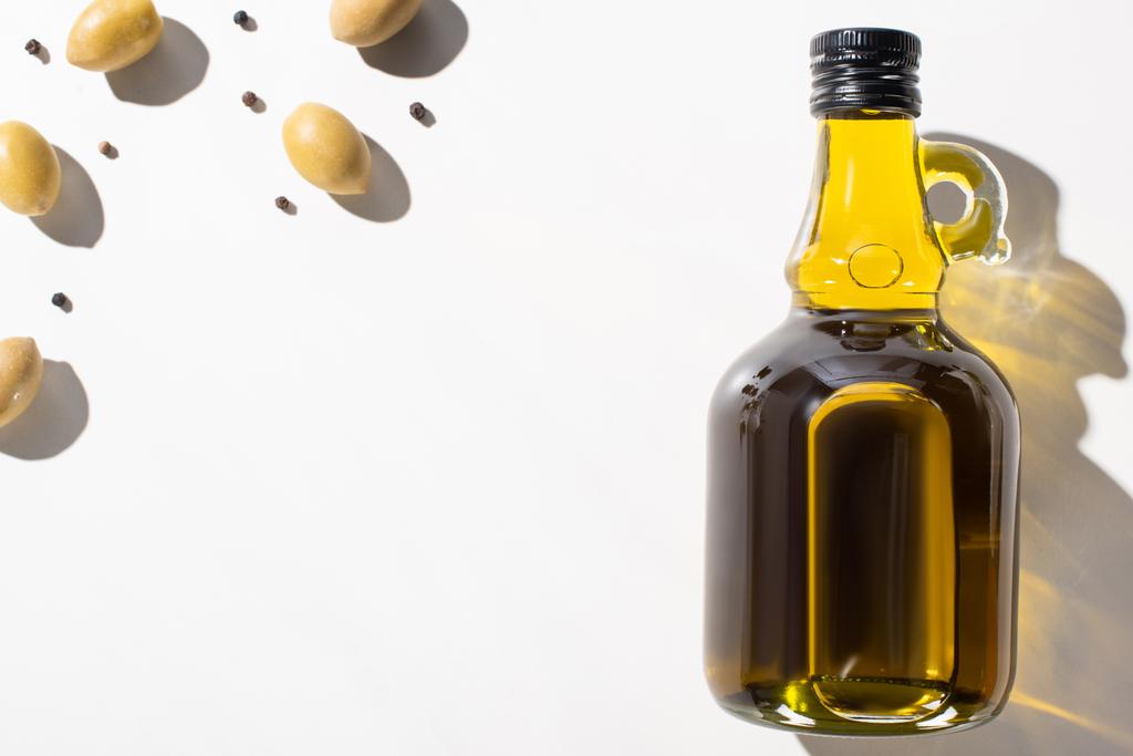Ansicht von Olivenöl in der Flasche in der Nähe von grünen Oliven und schwarzem Pfeffer auf weißem Hintergrund - Foto, Bild