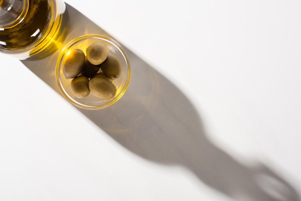 Ansicht von Olivenöl in Flasche in der Nähe von grünen Oliven in Schale im Schatten auf weißem Hintergrund - Foto, Bild