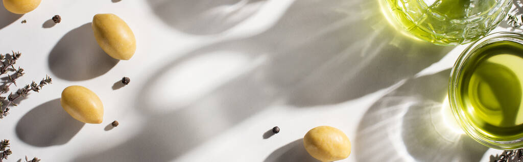 vista dall'alto dell'olio d'oliva in bottiglia e ciotola vicino alle erbe, olive verdi e pepe nero su fondo bianco con ombra, orientamento panoramico  - Foto, immagini