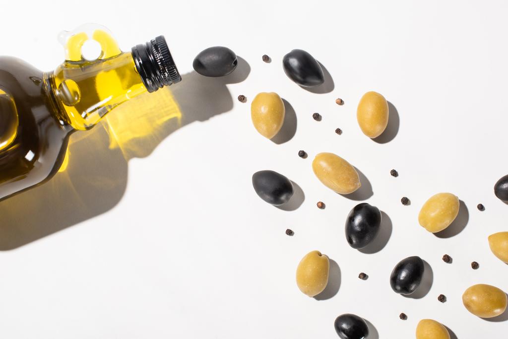 Ansicht von Olivenöl in der Flasche in der Nähe von grünen und schwarzen Oliven und schwarzem Pfeffer auf weißem Hintergrund mit Schatten - Foto, Bild
