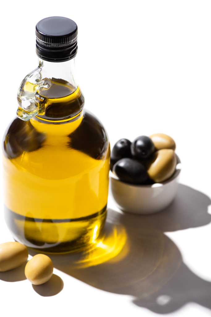olivový olej v láhvi v blízkosti zelené a černé olivy v misce na bílém pozadí - Fotografie, Obrázek