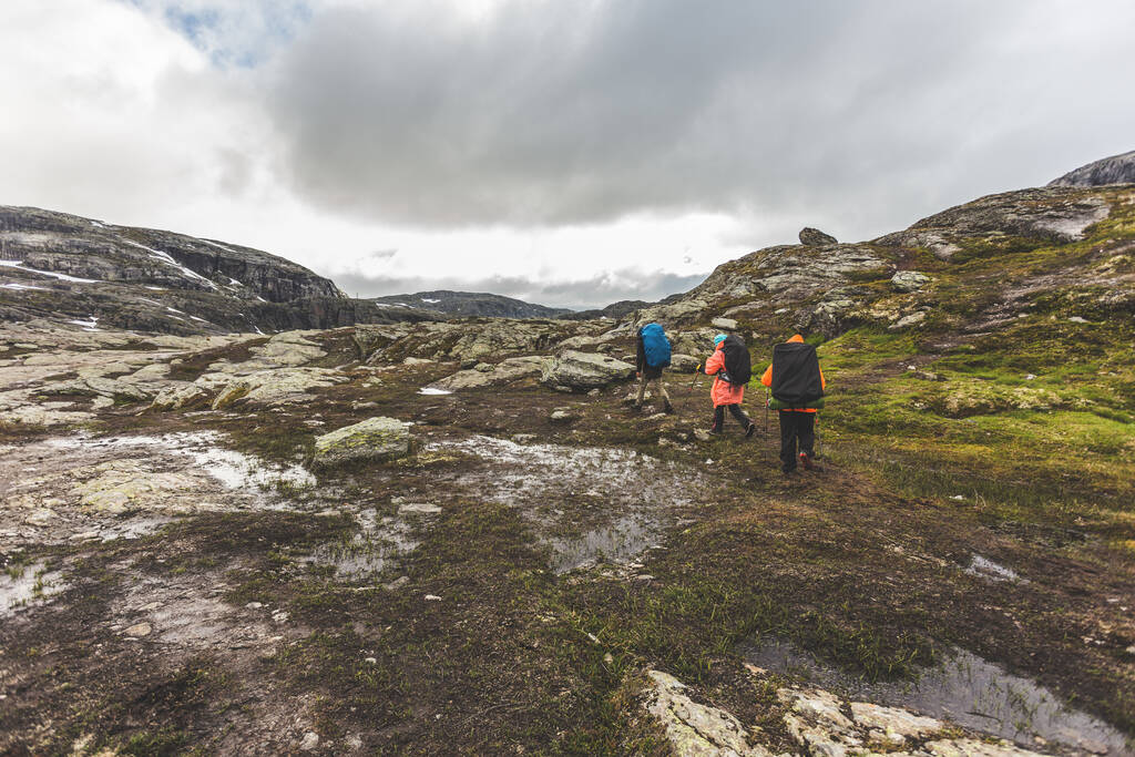 Touristen mit Rucksäcken wandern über eine felsige Ebene zum berühmten Wahrzeichen - dem Trollzungenfelsen oder Trolltunga. Kalter und windiger Sommer in Norwegen.  - Foto, Bild