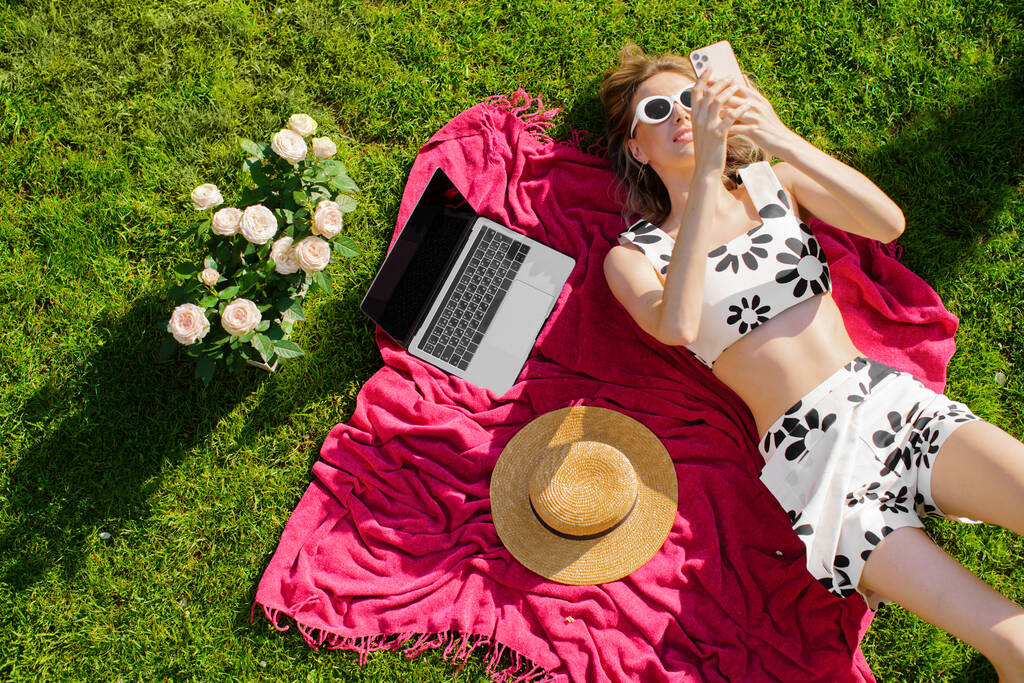 Junge hübsche Frau mit Sonnenbrille liegt auf dem Rasen und surft mit Laptop, Hut und Rosen im Internet, Foto für Blog oder Anzeige - Foto, Bild