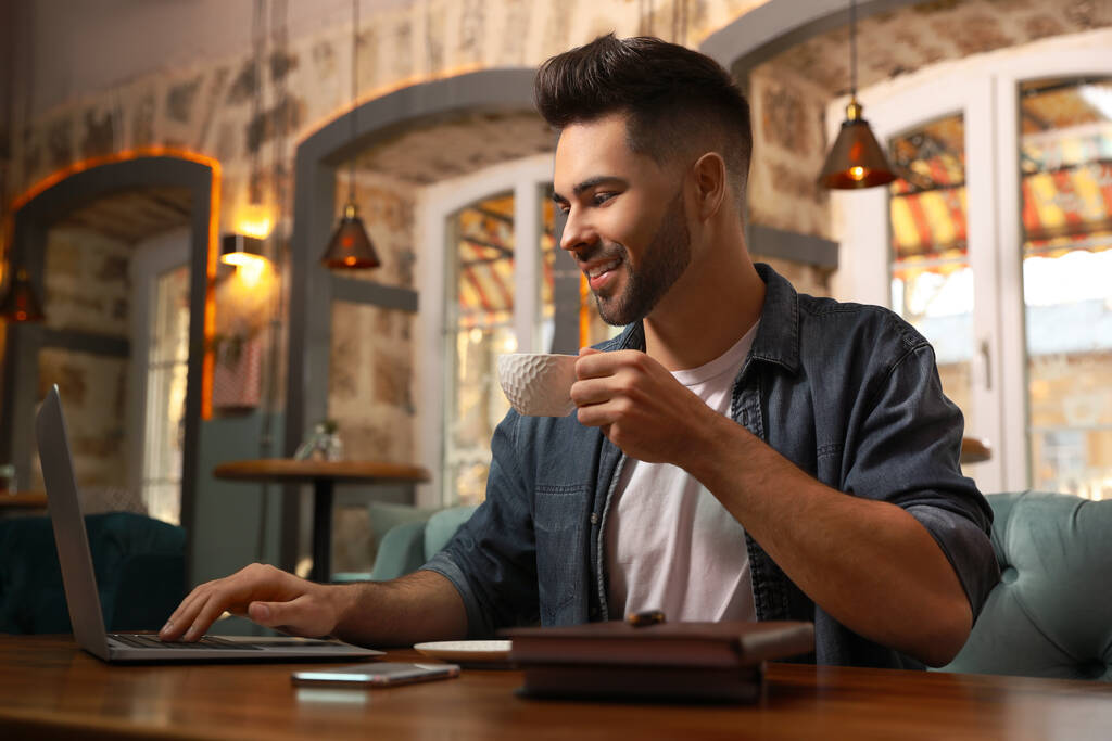 Молодой блогер с ноутбуком пьет кофе за столом в кафе
 - Фото, изображение